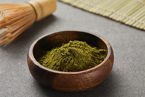 selective focus of bamboo whisk and green matcha powder in wooden bowl on grey table with copy space