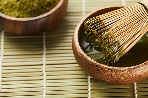 selective focus of green matcha powder and bamboo whisk in wooden bowl with tea