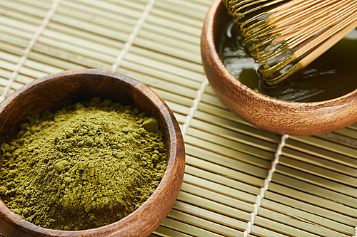 selective focus of green matcha powder and bamboo whisk in wooden bowl with tea on table mat