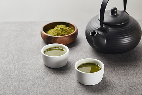 selective focus of green matcha powder in wooden bowl near 홍차pot and white cups with tea on grey stone table