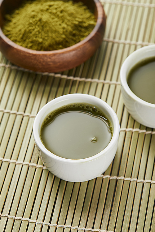 selective focus of green matcha powder in wooden bowl near white cups with tea on bamboo table mat