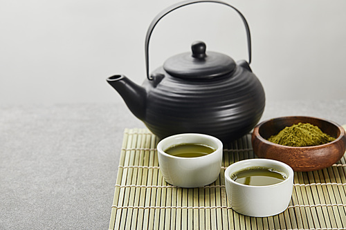 selective focus of green matcha powder in wooden bowl near 홍차pot and cups with tea on bamboo table mat