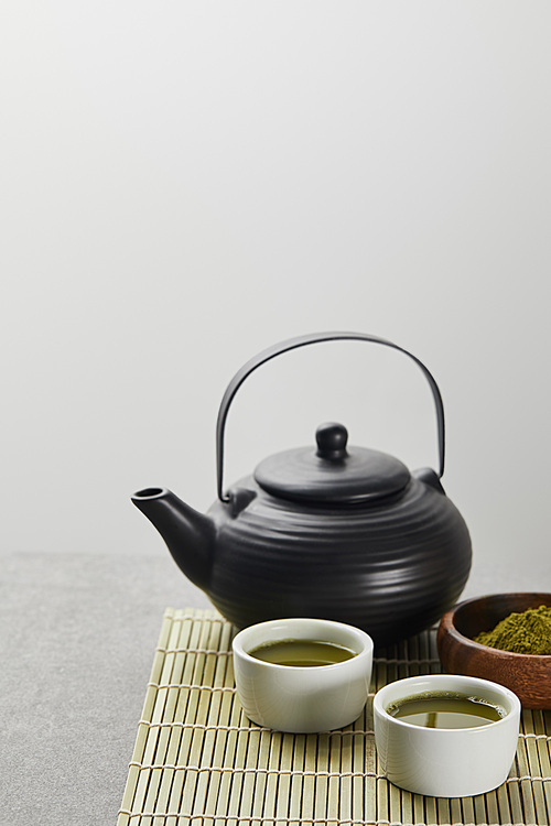 selective focus of green matcha powder in wooden bowl near 홍차pot and cups with tea on bamboo table mat