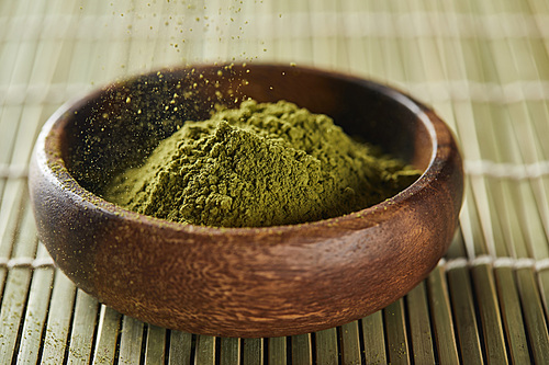 selective focus of green matcha powder pouring in wooden bowl on bamboo table mat