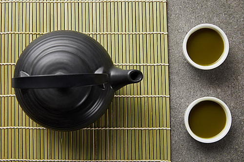 top view of white cups with green matcha tea near black teapot on bamboo table mat