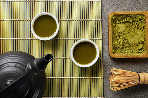 top view of white cups with green tea near 홍차pot on bamboo table mat with matcha powder on wooden board