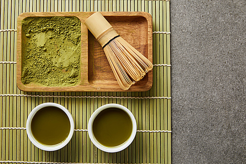 top view of green matcha powder with bamboo whisk on wooden board near white cups with green tea on table mat
