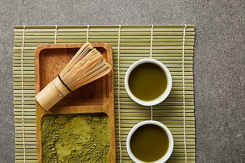 top view of green matcha powder with bamboo whisk on wooden board near white cups with green tea on table mat