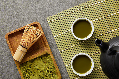 top view of green matcha powder with bamboo whisk in wooden board near teapot and white cups with green tea on table mat