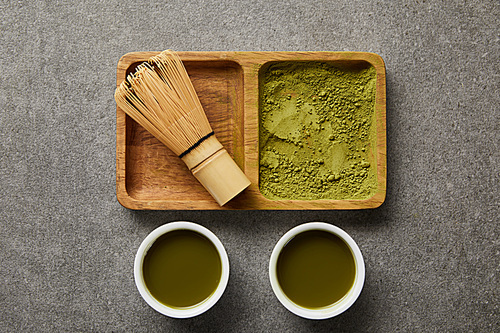top view of bamboo whisk and matcha powder on wooden bowl near white cups with green tea