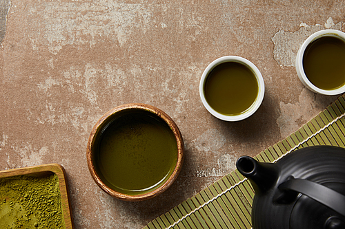 top view of matcha powder, bowl and cups with green tea on aged surface near bamboo table mat and 홍차pot