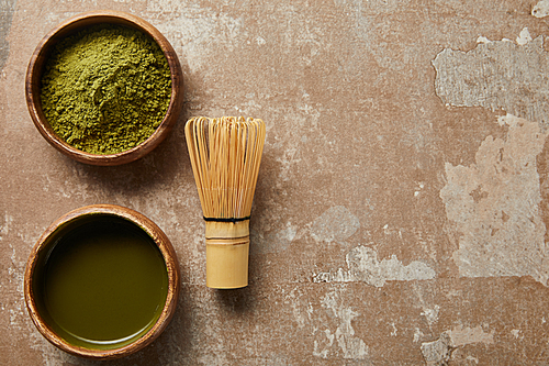 top view of matcha tea and green powder in wooden bowl with bamboo whisk on aged surface