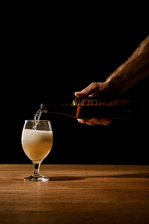 partial view of man pouring beer from bottle in glass on wooden table isolated on black