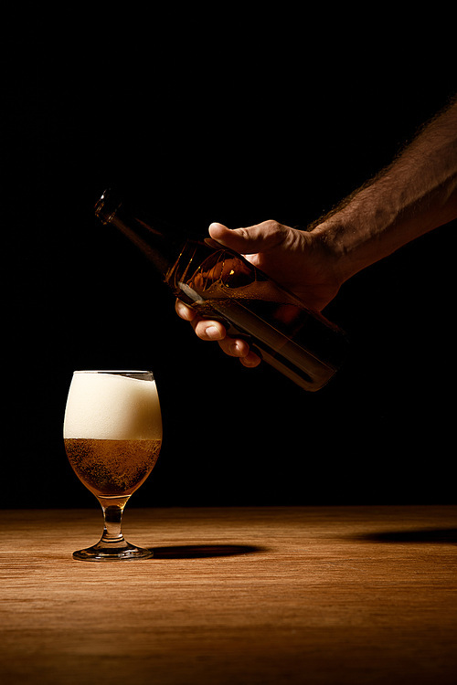 cropped view of man holding bottle near glass with beer and foam on wooden table isolated on black
