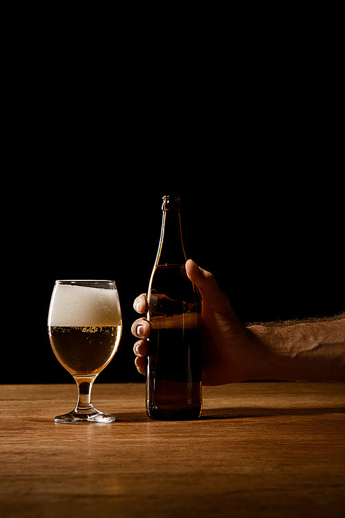 partial view of man holding bottle near glass with beer and foam on wooden table isolated on black