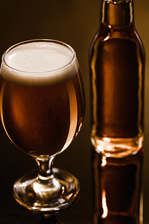 close up view of beer with foam in glass near bottle on dark background with lighting