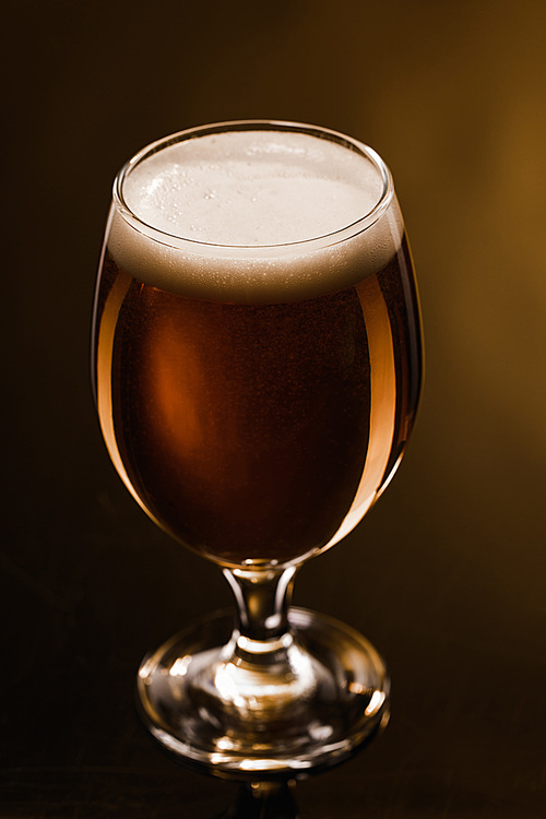 close up view of beer with foam in glass on dark background with lighting