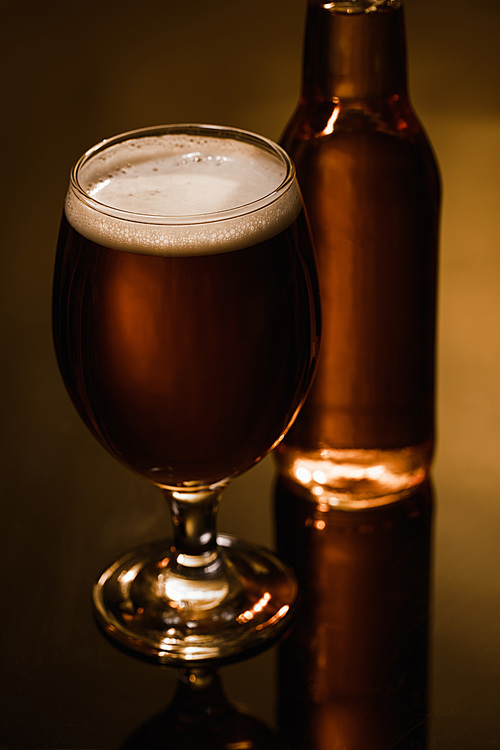 selective focus of beer with foam in glass near bottle on dark background with lighting