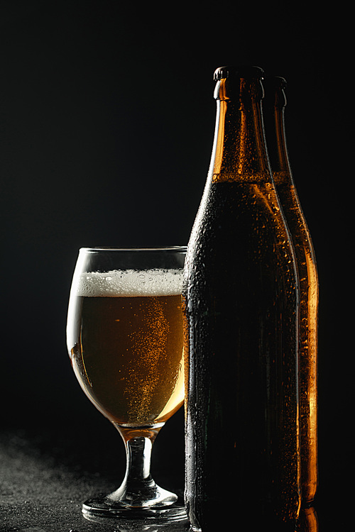 wet bottles of beer with drops near glass with beer and foam isolated on black