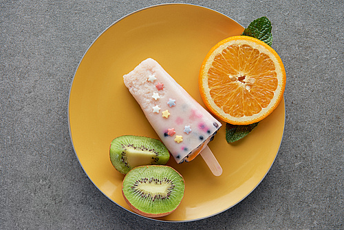 top view of delicious popsicle with slices of orange and kiwi on yellow plate on grey