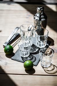 high angle view of fresh limes, empty glasses, tongs and shaker on wooden table