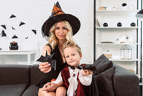 mother and little son in halloween costumes showing black paper bats on sofa at home