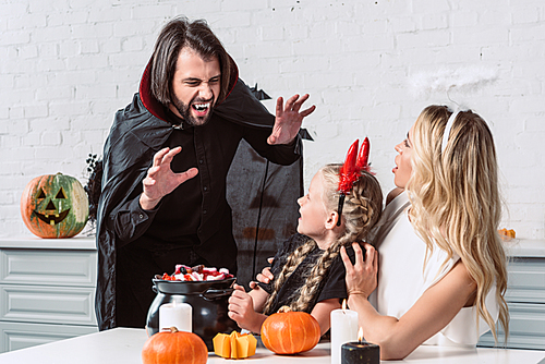 portrait of parents and daughter in halloween costumes at table with treats in black pot in kitchen at home