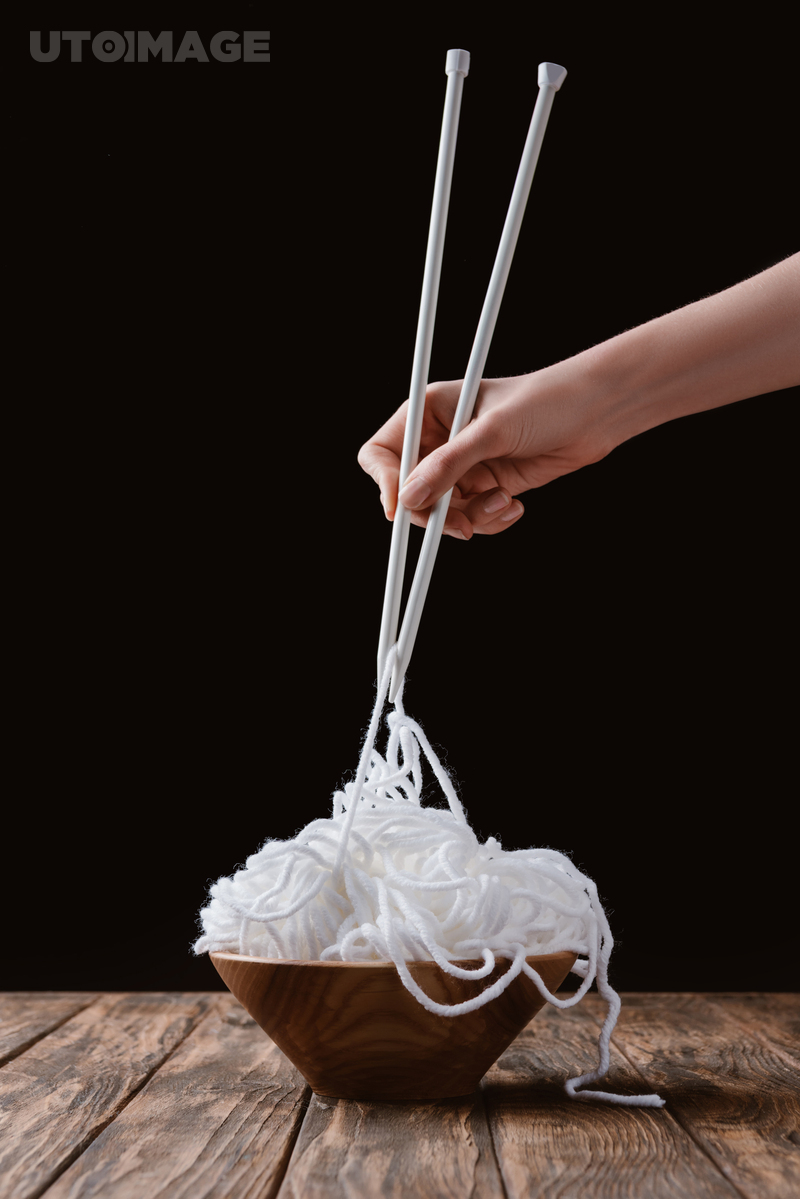 유토이미지 | cropped shot of woman holding white yarn with knitting needles like noodles on wooden tabletop on black background