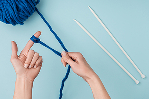 partial view of woman with blue yarn and white knitting needles knitting on blue backdrop
