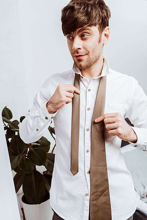 happy businessman in white shirt tying neck tie at home