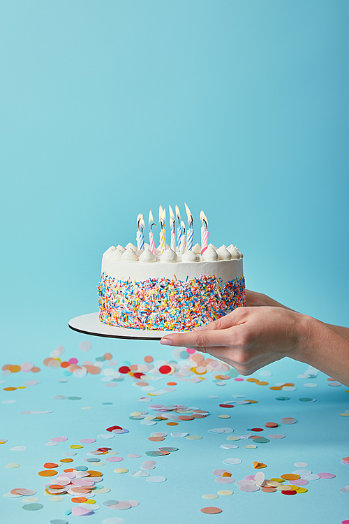 Cropped view of woman holding delicious birthday cake with candles on blue background with confetti