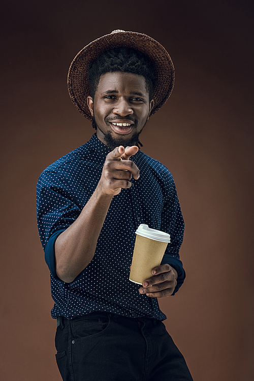african american man with disposable coffee cup pointing on camera isolated on brown