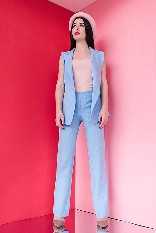 gorgeous young woman in stylish suit and hat looking away while standing on pink