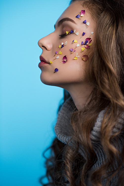 side view of young woman with flowers on face in sweater isolated on blue