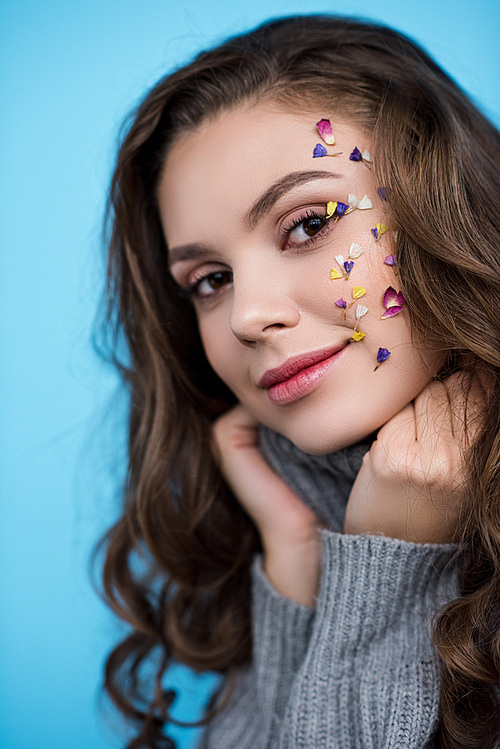 smiling young woman with flowers on face in turtleneck sweater  isolated on blue