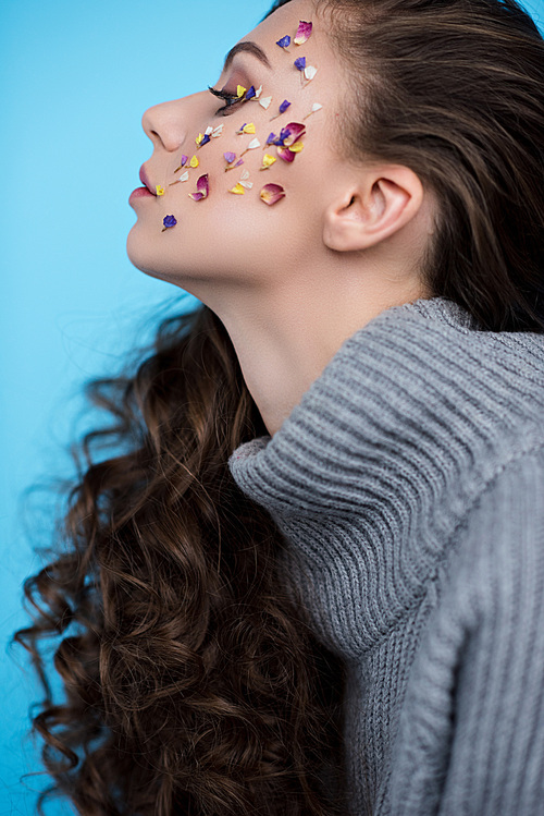 side view of young woman with flowers on face in warm sweater isolated on blue