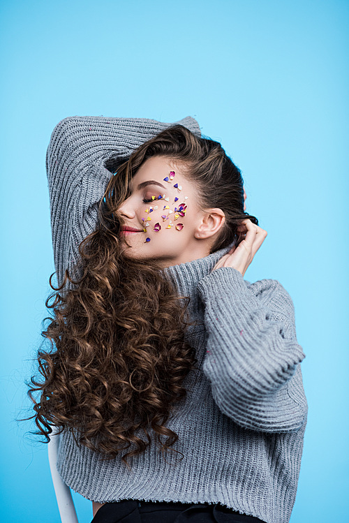 happy young woman with flower petals on face in sweater isolated on blue