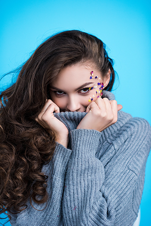 smiling young woman with flowers on face in stylish turtleneck sweater isolated on blue