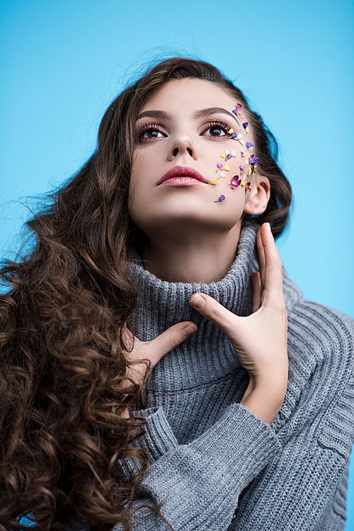 stylish woman in grey knitted turtleneck sweater with flowers on face looking up while posing isolated on blue