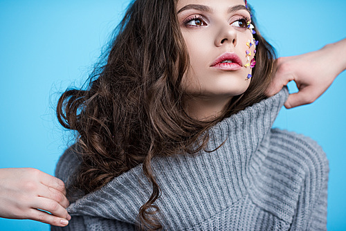 woman posing in warm sweater with flowers on face while people trying to stretch its neck isolated on blue