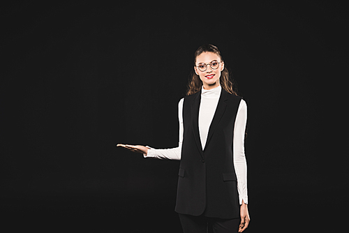 beautiful brunette woman in eyeglasses showing copy space and smiling at camera isolated on black