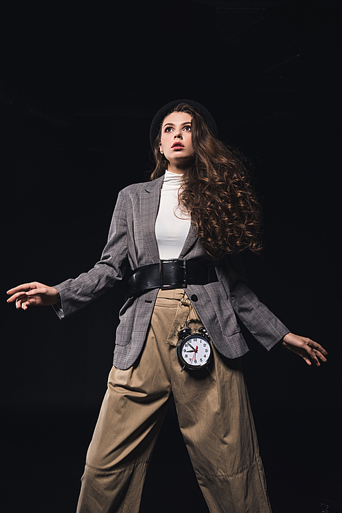 low angle view of fashionable shocked young woman with clock looking away isolated on black