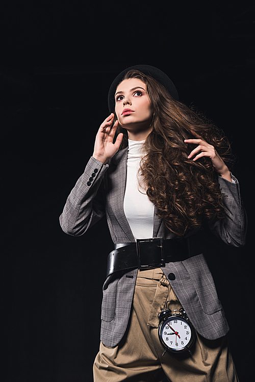 low angle view of fashionable pensive young woman with clock looking away isolated on black