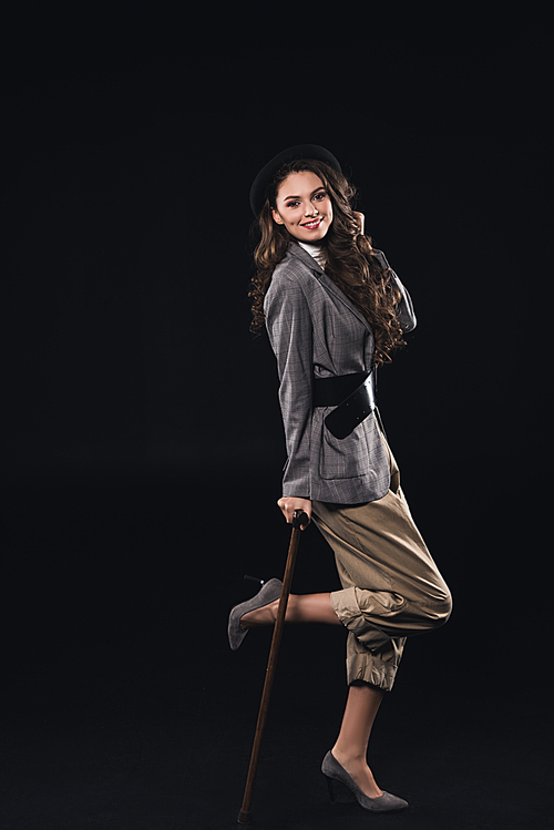 fashionable brunette woman posing with walking stick and smiling at camera isolated on black