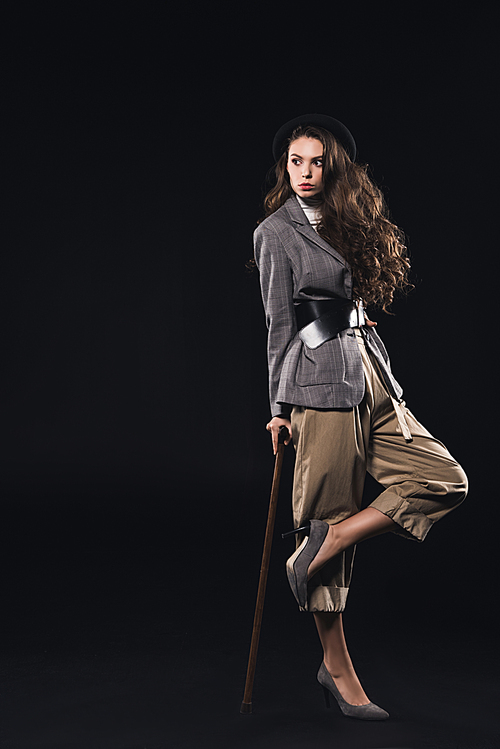 stylish young woman posing with walking stick and looking away isolated on black