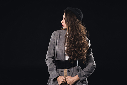 beautiful brunette woman in jacket with black belt and hat looking away isolated on black