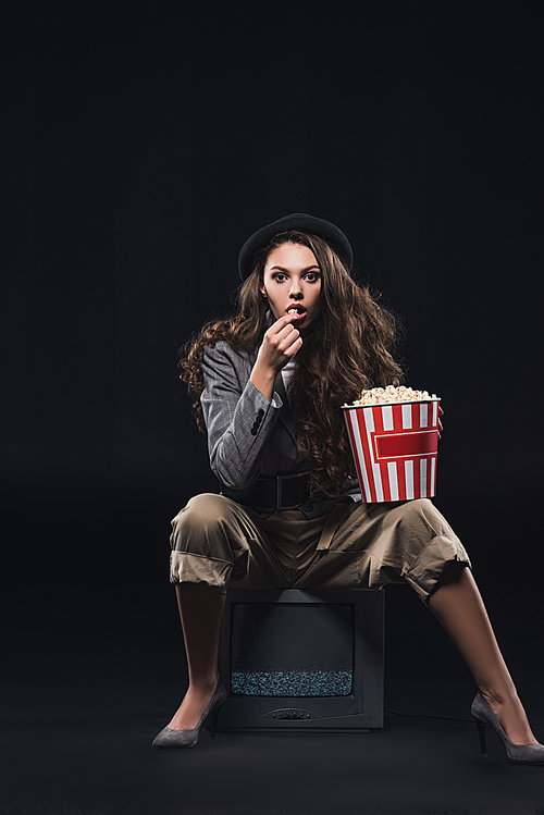 shocked young woman eating popcorn while sitting on tv and  on black