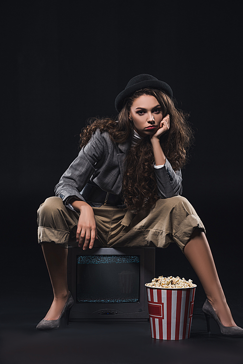 serious stylish young woman sitting on tv with popcorn and  on black