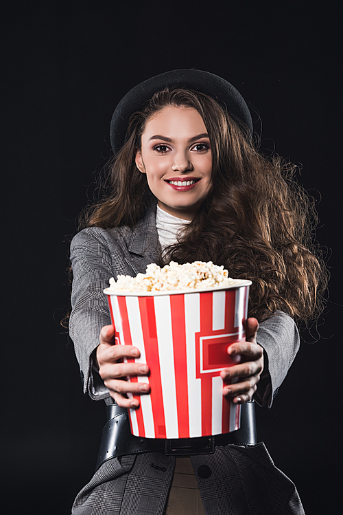 beautiful elegant young woman holding popcorn and smiling at camera isolated on black