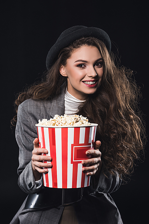 beautiful stylish young woman holding popcorn and smiling at camera isolated on black